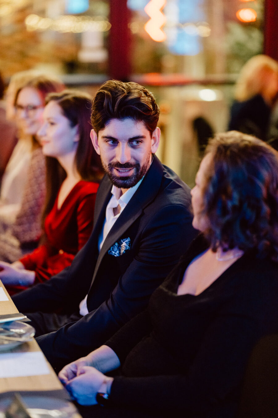 Mężczyzna w garniturze siedzi przy stole, rozmawiając z kobietą obok niego. Inne osoby siedzą w pobliżu, a tło zawiera ciepłe, rozmyte światła, tworząc przytulną atmosferę.