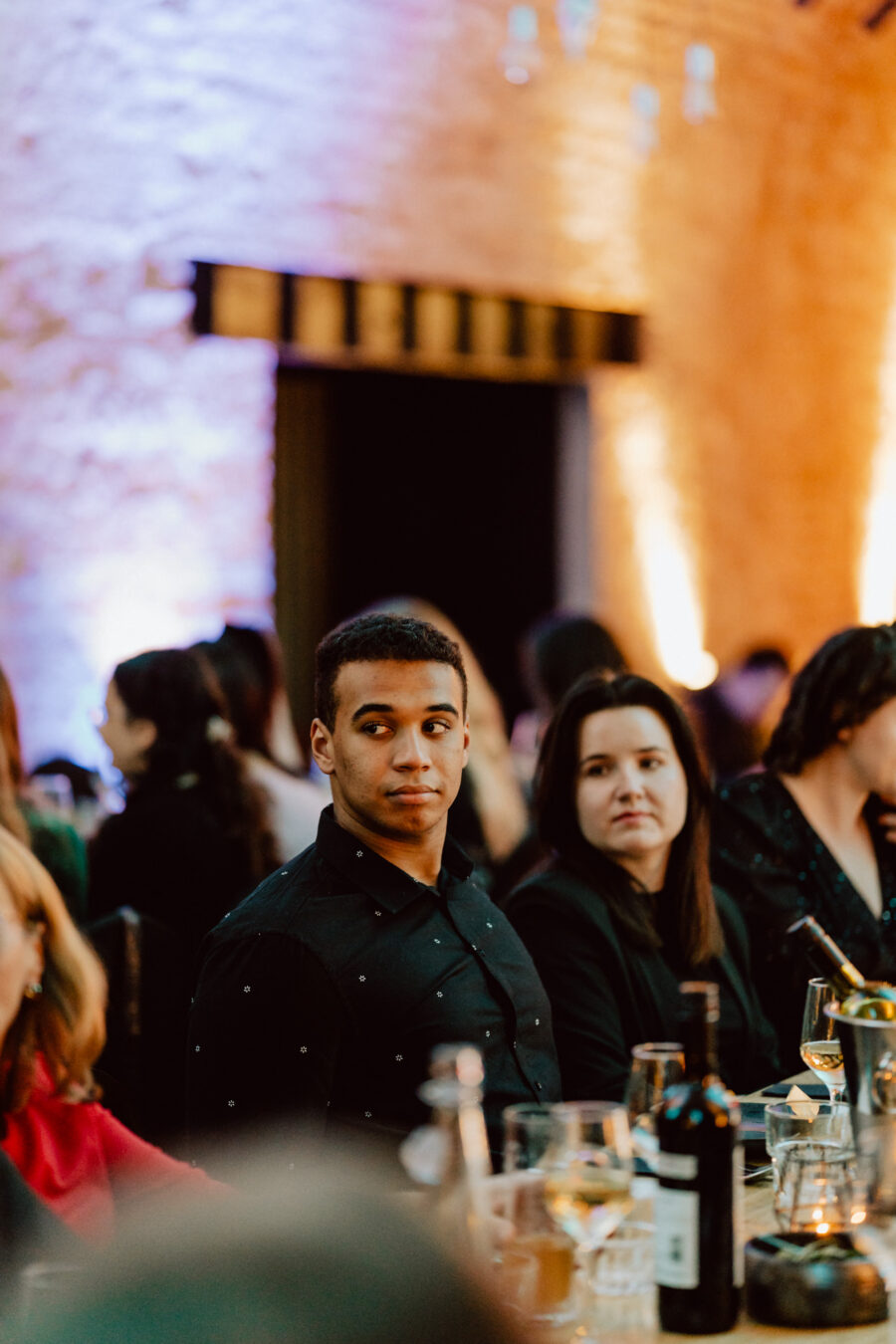 Mężczyzna i kobieta siedzą przy stole z kieliszkami i butelkami wina, otoczeni innymi ludźmi w ciepło oświetlonym pokoju o ceglanych ścianach. Mężczyzna patrzy w lewo, podczas gdy kobieta patrzy przed siebie.