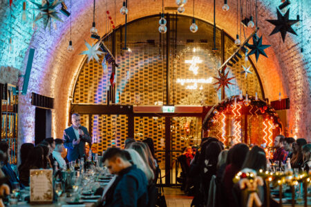 fototograf na impreze firmowa browary warszawskie wigilia 024 Grupa ludzi siedzi przy udekorowanych stołach w świątecznym, łukowatym miejscu z cegły z wiszącymi gwiazdami i światłami, słuchając mówcy w garniturze zwracającego się do publiczności z przodu.