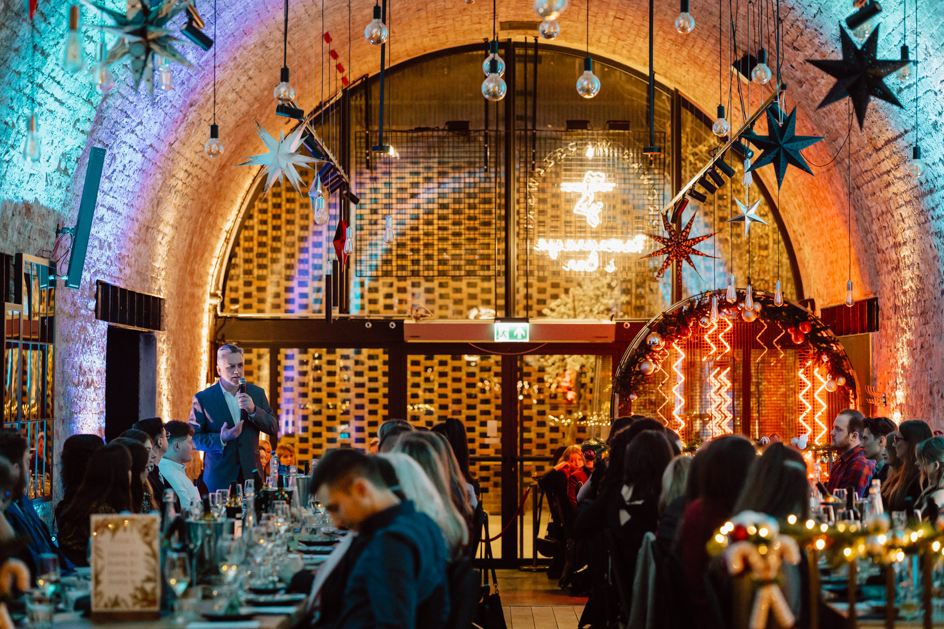 A group of people sit at decorated tables in a festive arched brick venue with hanging stars and lights, listening to a speaker in a suit addressing the audience up front.