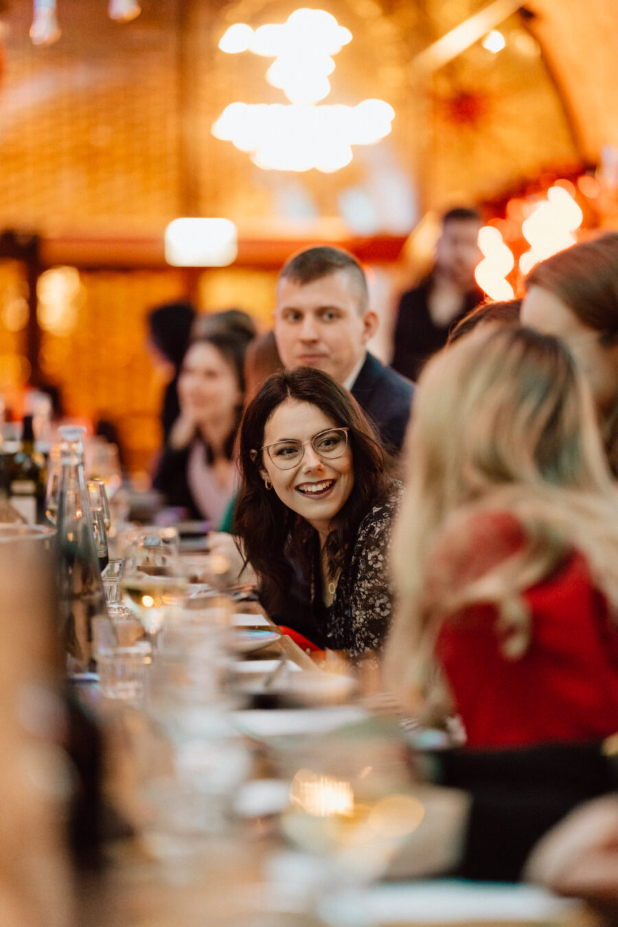 Kobieta w okularach uśmiecha się promiennie, siedząc przy długim, świątecznym stole otoczonym ludźmi w ciepło oświetlonej, tętniącej życiem restauracji lub przestrzeni eventowej.