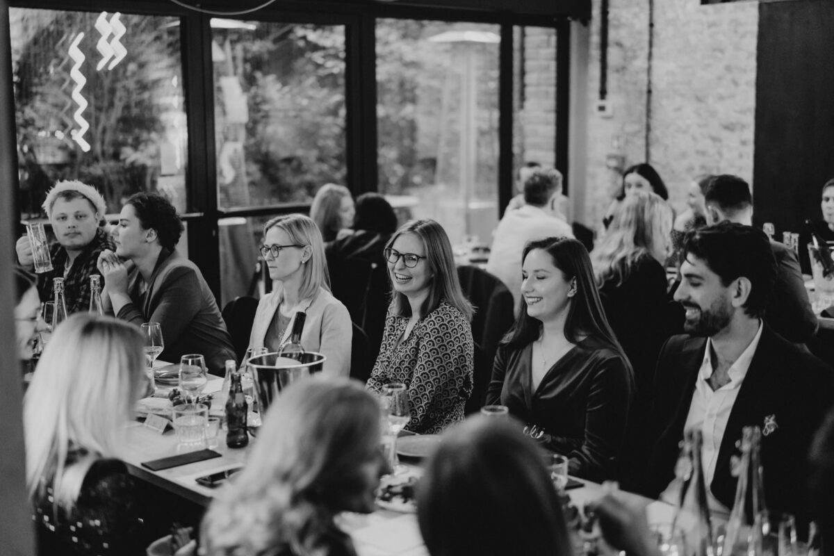 Grupa ludzi siedzi przy długim stole w restauracji, zaangażowana w rozmowę i śmiech podczas spotkania towarzyskiego. Atmosfera jest zrelaksowana i ożywiona.