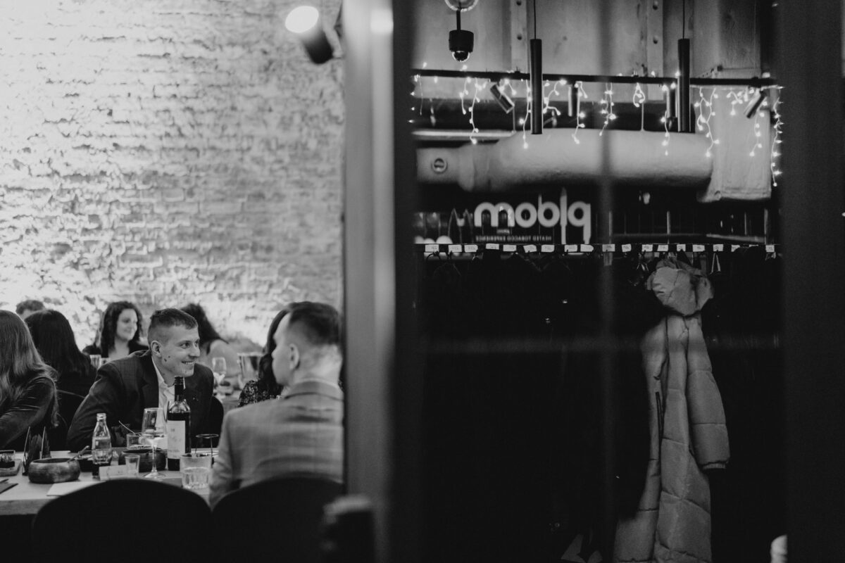 Czarno-białe zdjęcie ludzi jedzących posiłek w restauracji, z dwoma mężczyznami rozmawiającymi przy stoliku na pierwszym planie. Płaszcze wiszą na wieszakach, a światła sznurkowe zdobią ścianę w tle.