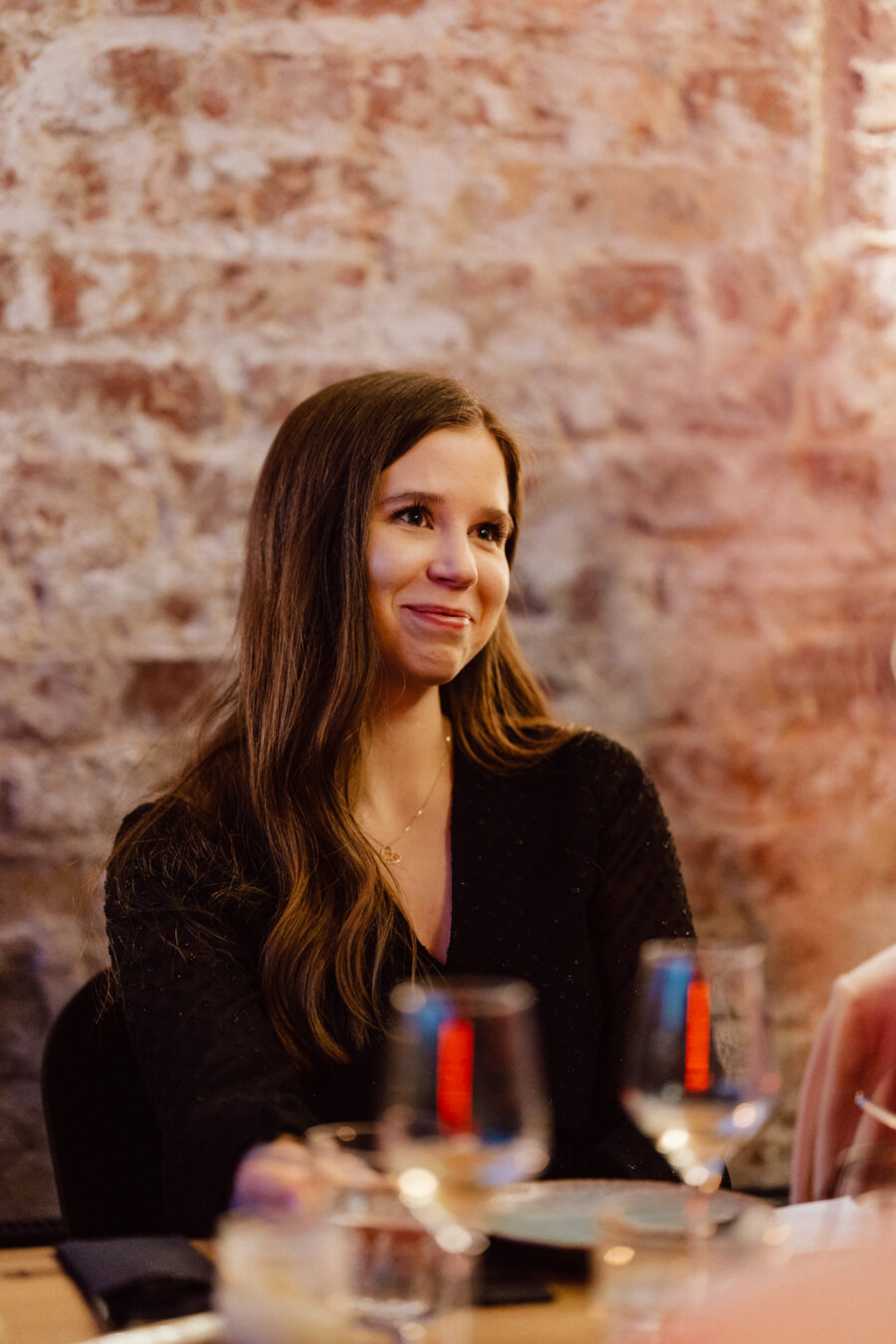 Młoda kobieta z długimi brązowymi włosami, ubrana w czarny top, siedzi przy stole przed ceglaną ścianą, uśmiechając się delikatnie. Na pierwszym planie znajdują się szklanki i zastawa stołowa.