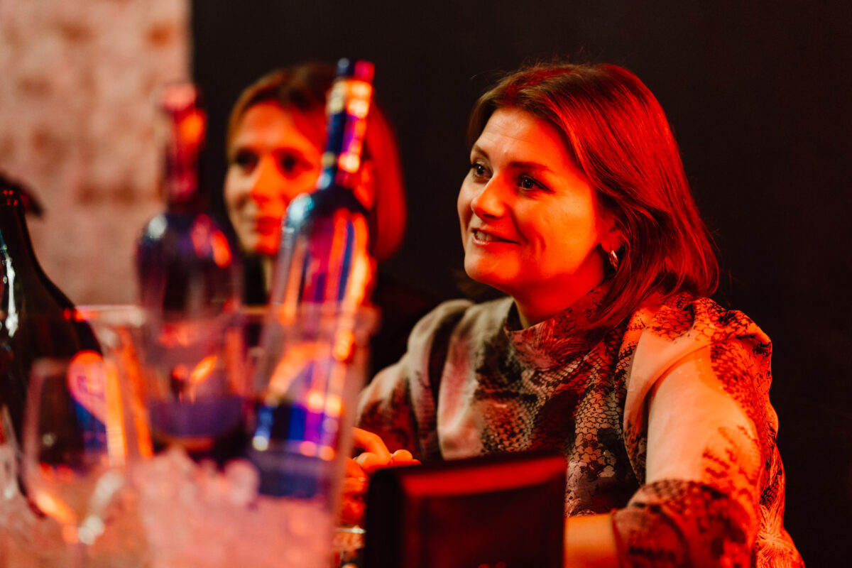 Dwie kobiety siedzą przy stole z butelkami i szklankami na pierwszym planie, jedna z nich uśmiecha się i patrzy w bok w ciepłym, pomarańczowym oświetleniu.