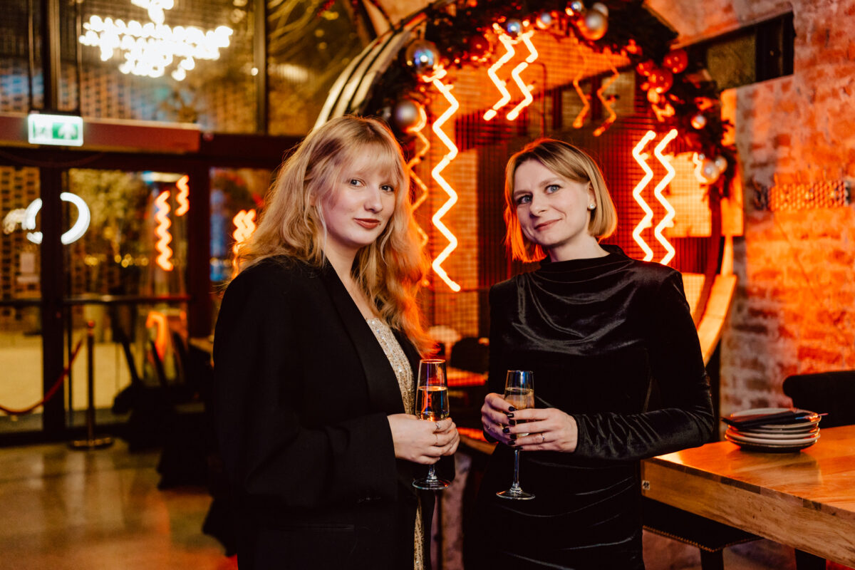 Dwie kobiety w eleganckich strojach wieczorowych stoją w eleganckim lokalu, trzymając kieliszki szampana. W tle widać neony i świąteczne dekoracje, tworząc ciepłą, uroczystą atmosferę.