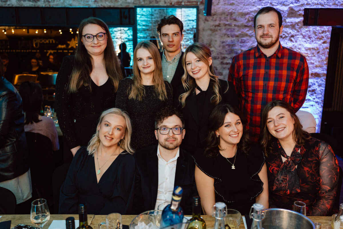 Grupa dziewięciu osób, ubranych w półformalne stroje, pozuje i uśmiecha się do siebie przy stole zastawionym napojami i butelkami wina w ciepło oświetlonej restauracji lub przestrzeni eventowej.