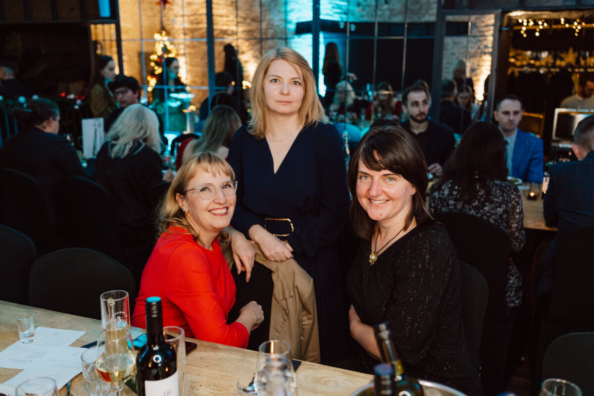 Trzy kobiety pozują i uśmiechają się do stołu podczas oficjalnego wydarzenia lub kolacji. Inni goście siedzą w tle. Pokój jest ciepło oświetlony świątecznymi dekoracjami i napojami na stole.