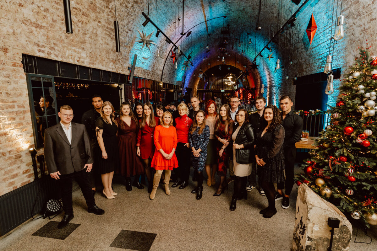 Grupa osób ubranych w formalne i półformalne stroje pozuje razem w świątecznym, łukowatym ceglanym korytarzu ozdobionym światłami i ornamentami, z choinką po prawej stronie.
