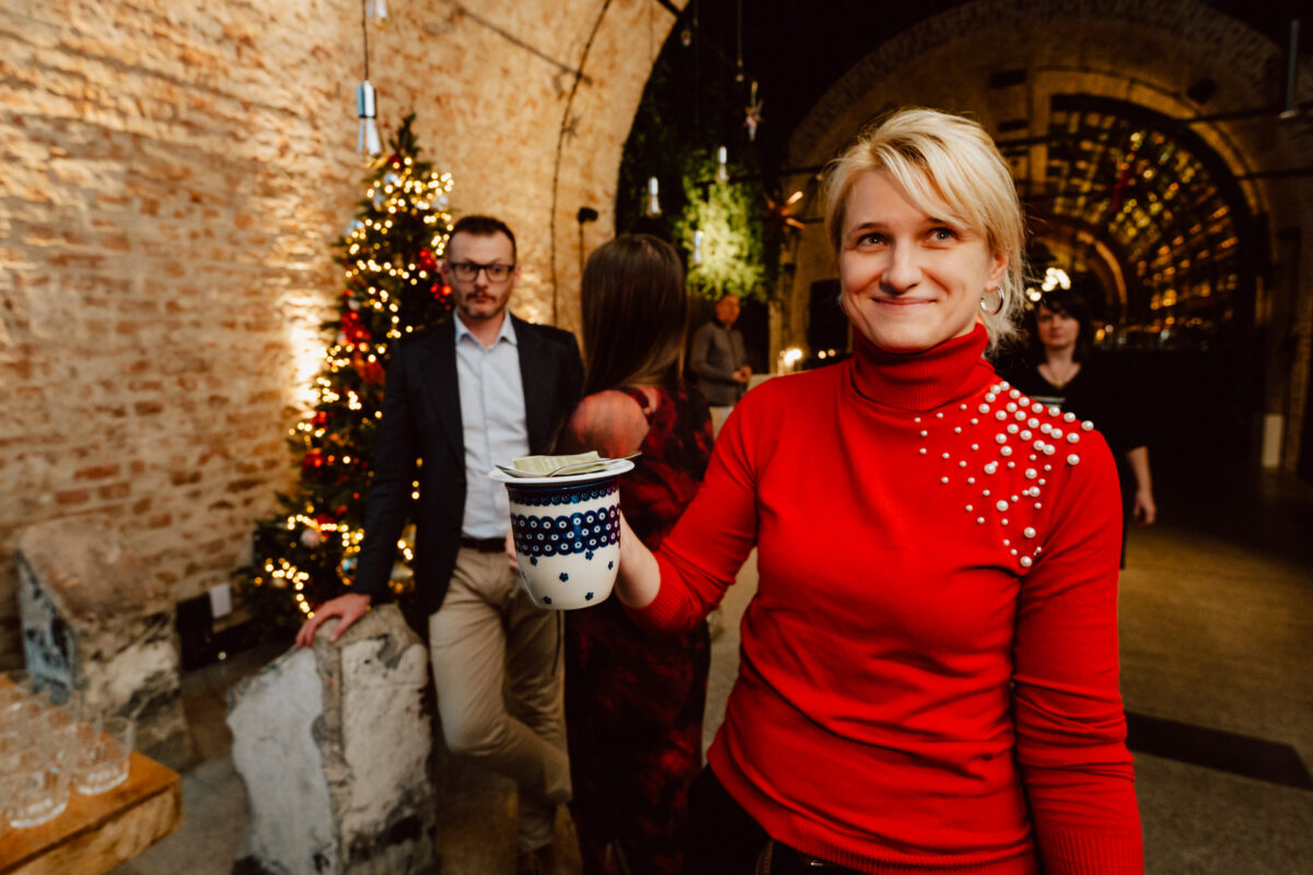 Kobieta w czerwonym swetrze z perłowymi zdobieniami uśmiecha się, trzymając kubek podczas świątecznego spotkania w domu, z choinką i innymi ludźmi w tle.