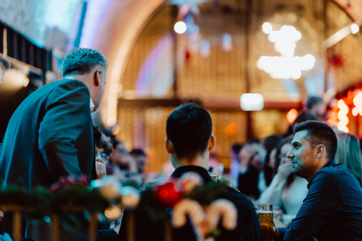 Grupa ludzi siedzi przy stole w tętniącej życiem, ciepło oświetlonej restauracji lub barze, rozmawiając i nawiązując kontakty towarzyskie. W tle widać miękkie, kolorowe światła i rozmazany żyrandol.