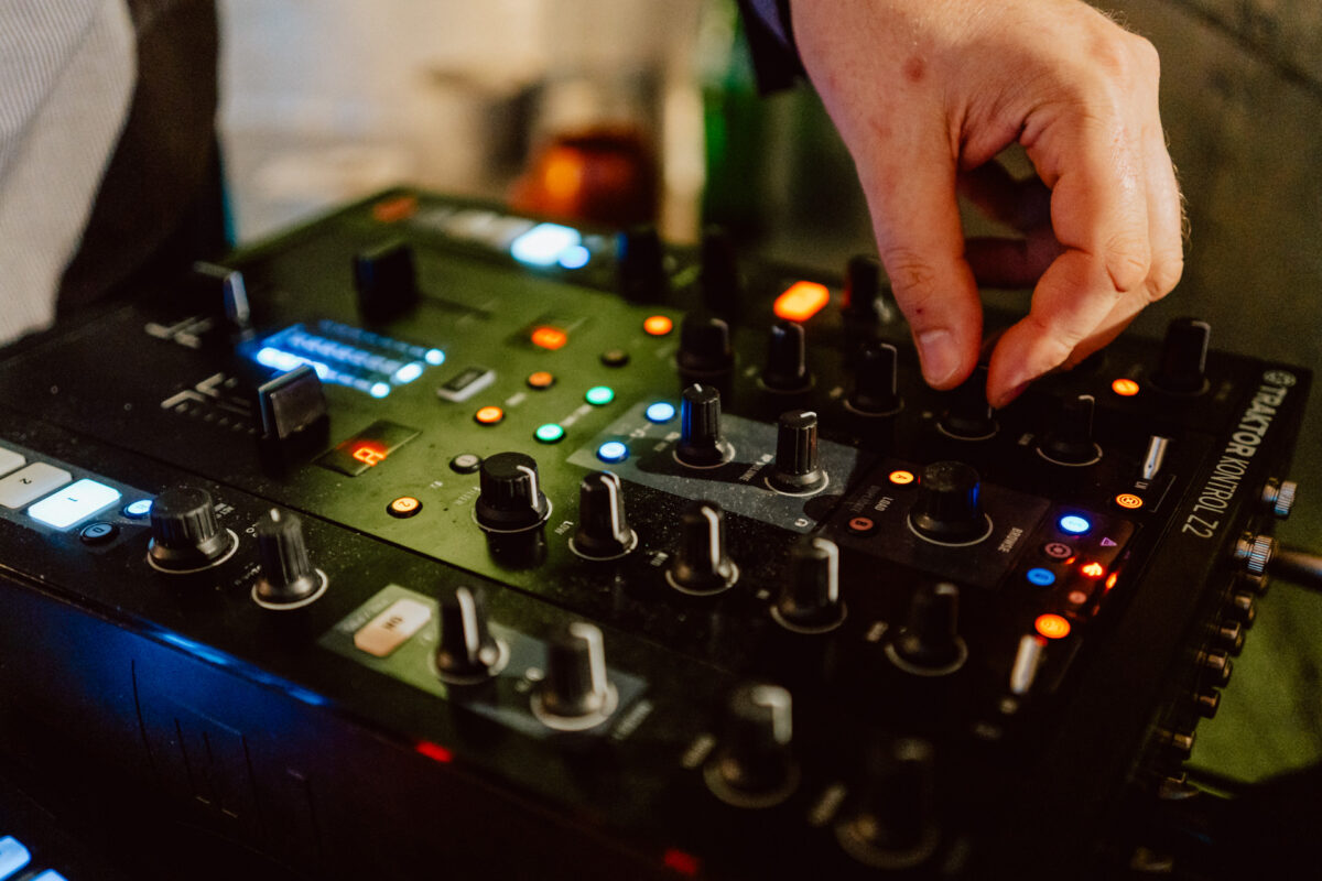 Zbliżenie dłoni osoby regulującej pokrętła i elementy sterujące na mikserze DJ-a, otoczonej kolorowymi światłami i przyciskami.