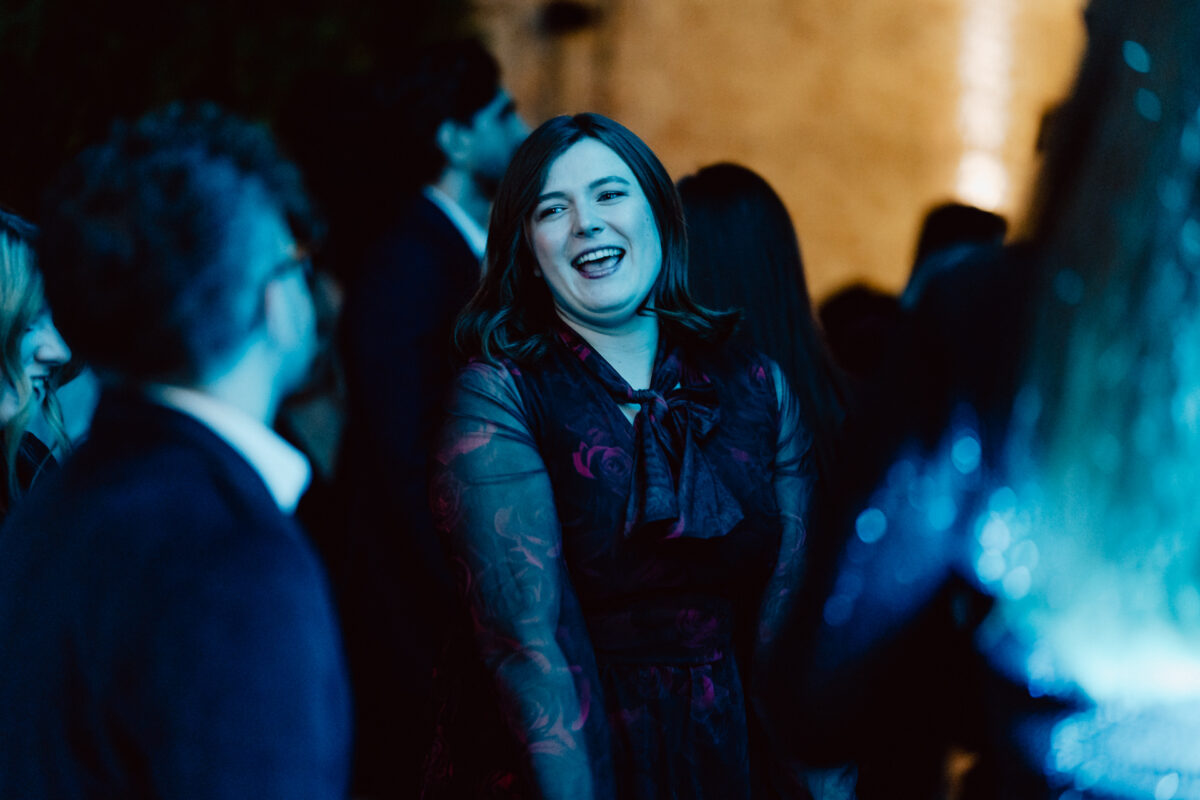 Kobieta w ciemnej, kwiecistej sukience uśmiecha się i śmieje podczas rozmowy z innymi na nocnej imprezie, oświetlonej niebieskim światłem. Ludzie stoją i rozmawiają wokół niej w ożywionej atmosferze.