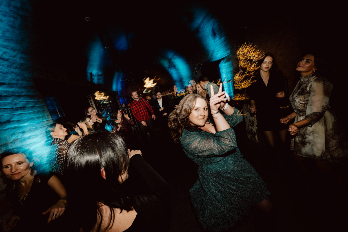 Tętniąca życiem grupa ludzi tańczy w słabo oświetlonym pokoju z niebieskim i żółtym oświetleniem. Kobieta na pierwszym planie trzyma telefon, prawdopodobnie robiąc selfie, podczas gdy inni tańczą i uśmiechają się w tle.