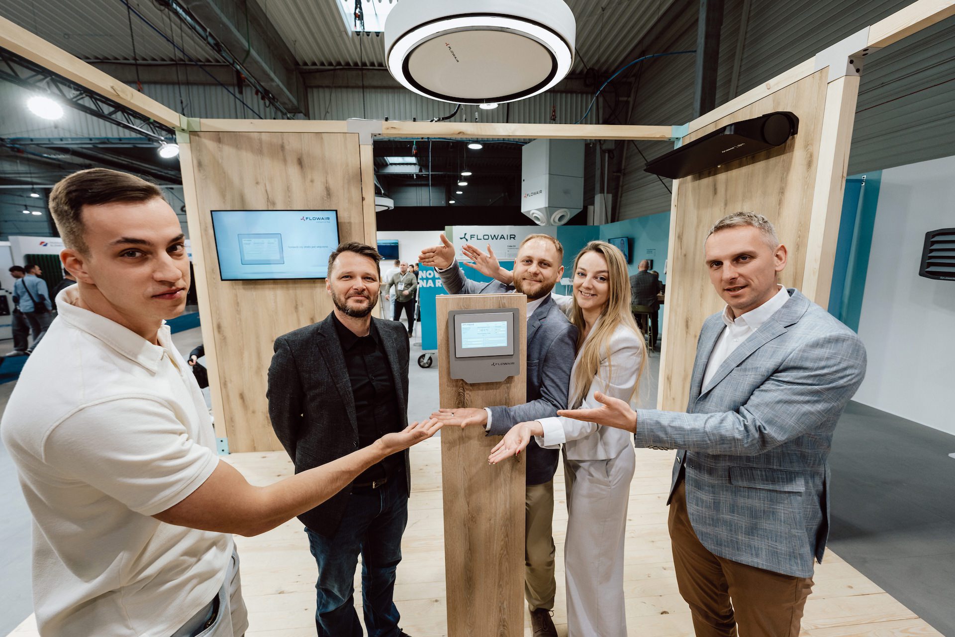 sesja employer branding na targach Pięć osób w strojach biznesowych stoi w pomieszczeniu, uśmiechając się i gestykulując w kierunku ekranu na stoisku wystawowym. Logo flowair jest widoczne na ekranach w tle, dzięki czemu ta fotografia targowa jest idealna do każdej fotorelacji z targów.