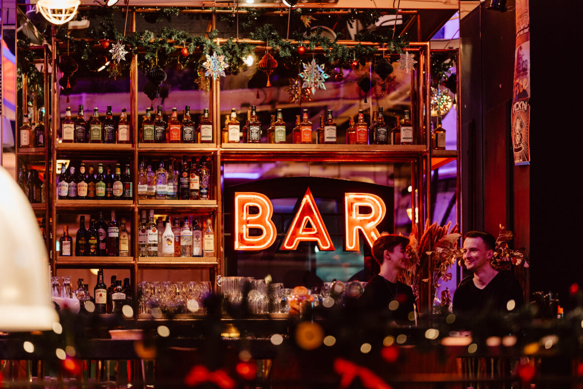 wigilia firmowa spoldzielnia koneser fotograf na impreze 001 Dwie osoby siedzą i rozmawiają przy barze, za którym znajdują się półki z butelkami alkoholu. W tle świeci jasny neon z napisem "BAR", a nad nim wiszą świąteczne dekoracje. Atmosfera jest ciepła i zachęcająca.
