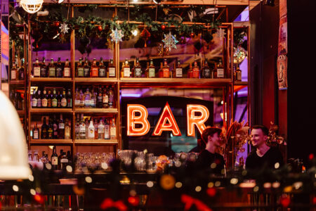 Dwie osoby siedzą i rozmawiają przy barze, za którym znajdują się półki z butelkami alkoholu. W tle świeci jasny neon z napisem "BAR", a nad nim wiszą świąteczne dekoracje. Atmosfera jest ciepła i zachęcająca.