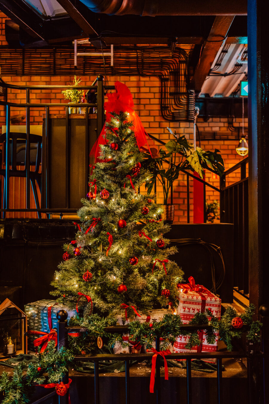wigilia firmowa spoldzielnia koneser fotograf na impreze 002 Mała choinka udekorowana czerwonymi ozdobami i światełkami stoi w pomieszczeniu w pobliżu zapakowanych prezentów i świątecznych girland, z ceglaną ścianą i roślinami w tle.