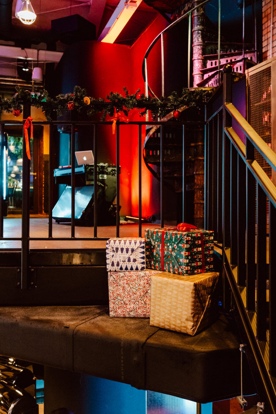 wigilia firmowa spoldzielnia koneser fotograf na impreze 003 Trzy zapakowane pudełka na prezenty stoją na podeście obok metalowych schodów ozdobionych girlandą i czerwonymi wstążkami w ciepło oświetlonym, świątecznym wnętrzu.