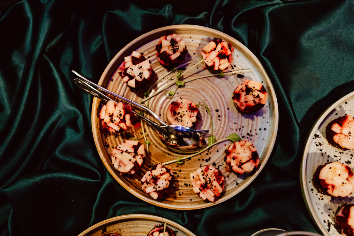 wigilia firmowa spoldzielnia koneser fotograf na impreze 004 Okrągły talerz z kilkoma małymi przekąskami pokrojonymi w kostkę, udekorowanymi ziołami i podawanymi srebrną łyżeczką na ciemnozielonym, jedwabistym obrusie.