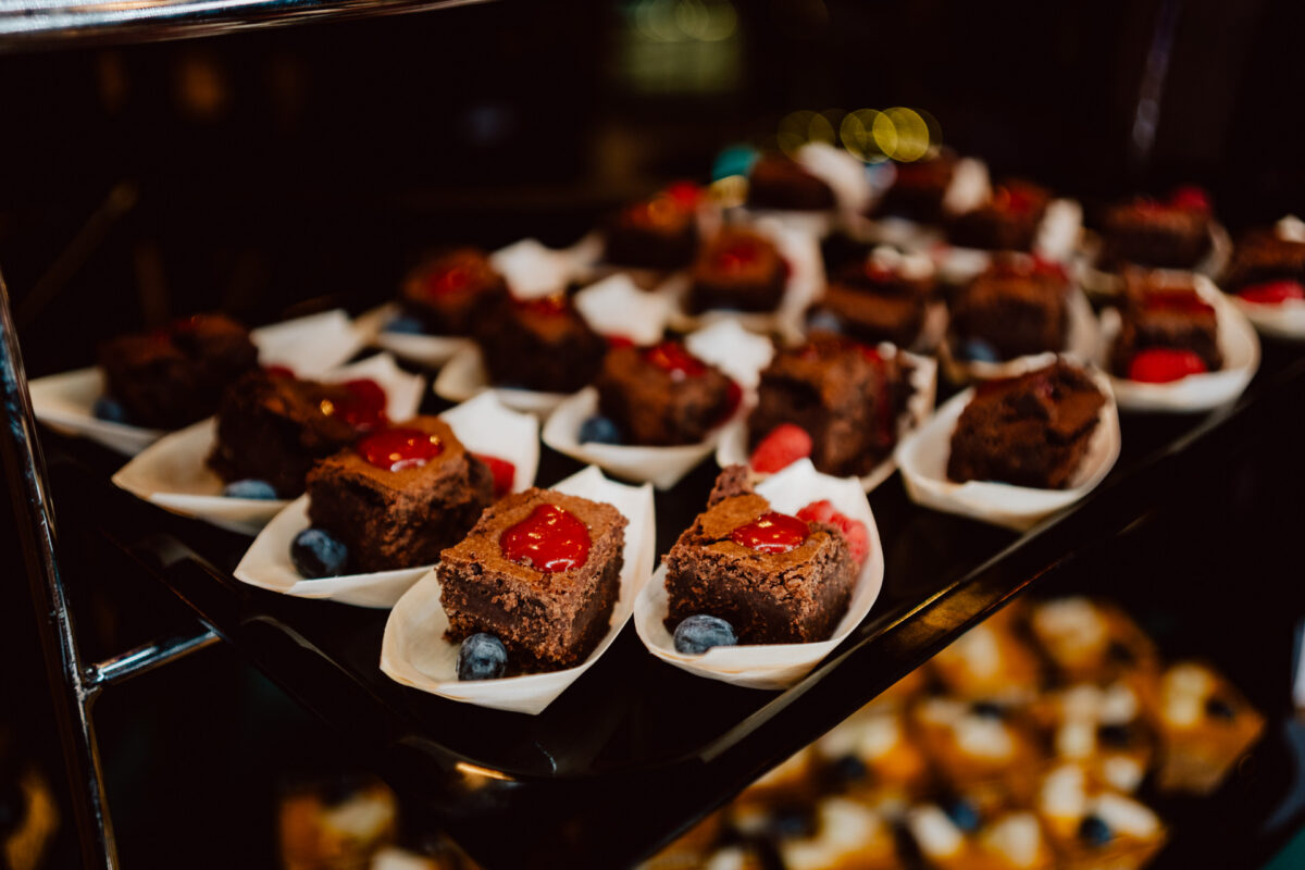 wigilia firmowa spoldzielnia koneser fotograf na impreze 005 Zbliżenie na czekoladowe brownies polane sosem z czerwonych owoców, podane na małych białych papierowych tackach z jagodami, ułożone na czarnej tacy.