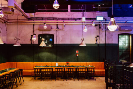 Nowoczesne wnętrze restauracji z pomarańczowymi wyściełanymi ławkami, długim drewnianym stołem z zielonymi serwetkami, czarnymi krzesłami, lampami wiszącymi w stylu industrialnym i odsłoniętymi rurami na ścianach.