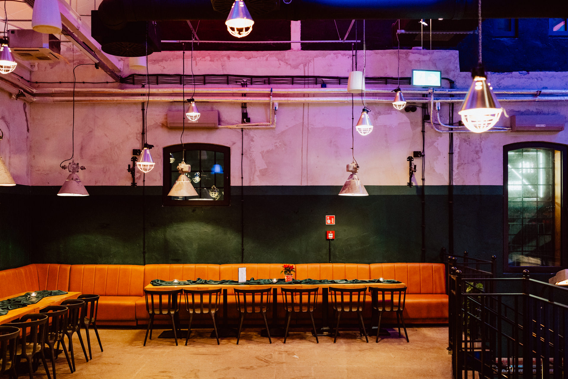 Nowoczesne wnętrze restauracji z pomarańczowymi wyściełanymi ławkami, długim drewnianym stołem z zielonymi serwetkami, czarnymi krzesłami, lampami wiszącymi w stylu industrialnym i odsłoniętymi rurami na ścianach.