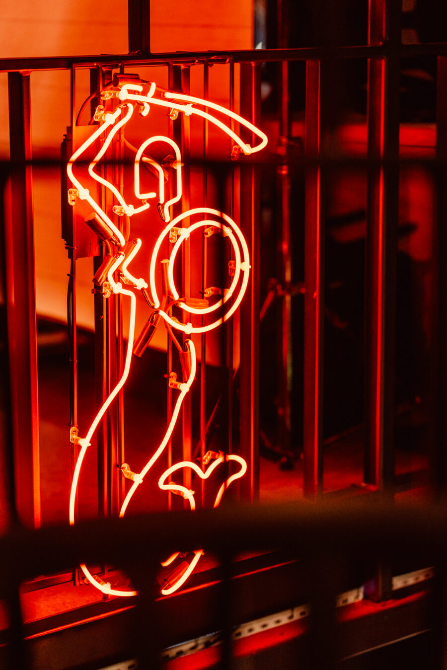 wigilia firmowa spoldzielnia koneser fotograf na impreze 009 Jasnoczerwony neon przedstawiający stylizowaną syrenę trzymającą miecz i tarczę, wyświetlany za pionowymi metalowymi prętami w słabo oświetlonym otoczeniu.