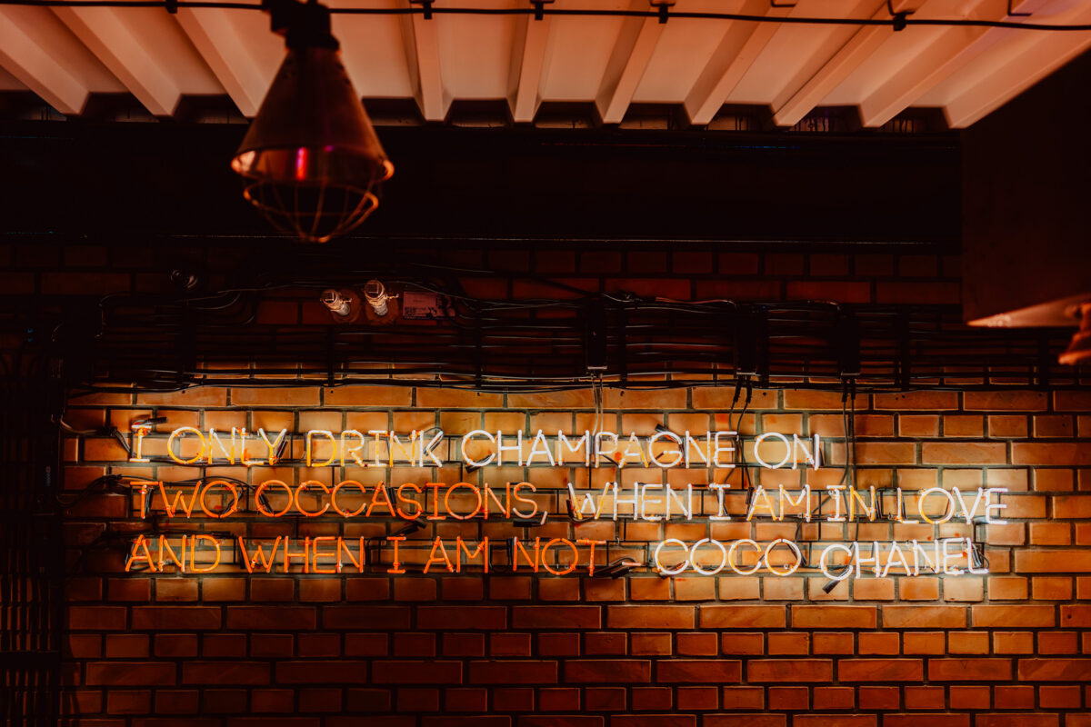 wigilia firmowa spoldzielnia koneser fotograf na impreze 011 Neonowy napis na ceglanej ścianie głosi: "Piję szampana tylko przy dwóch okazjach, kiedy jestem zakochana i kiedy nie jestem - Coco Chanel". Z sufitu powyżej zwisają lampy przemysłowe.