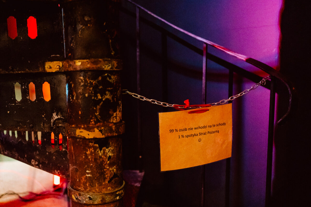 wigilia firmowa spoldzielnia koneser fotograf na impreze 013 Zardzewiałe spiralne schody są zablokowane łańcuchem z żółtym napisem w języku polskim. Tło jest słabo oświetlone czerwonym i fioletowym światłem.