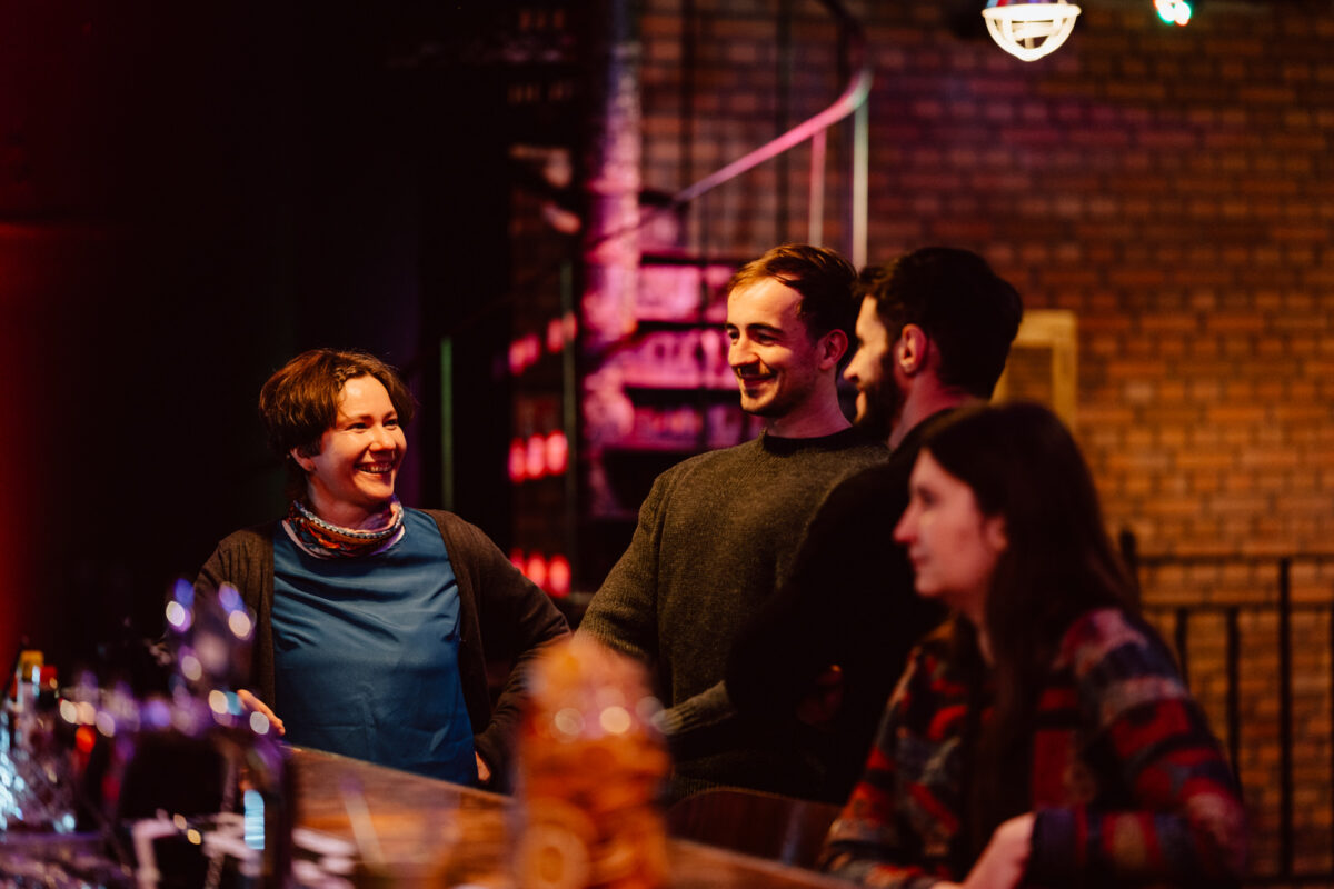 wigilia firmowa spoldzielnia koneser fotograf na impreze 016 Cztery osoby siedzą przy barze, uśmiechając się do siebie i rozmawiając. W tle znajdują się odsłonięte ceglane ściany i ciepłe oświetlenie, tworząc przytulną, relaksującą atmosferę. Na barze widoczne są różne napoje i szklanki.