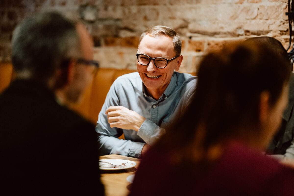 wigilia firmowa spoldzielnia koneser fotograf na impreze 017 Mężczyzna w okularach i jasnoniebieskiej koszuli siedzi przy stoliku, uśmiechając się i rozmawiając z innymi w ciepło oświetlonej, rustykalnej restauracji. Trzy inne osoby są częściowo widoczne na pierwszym planie.