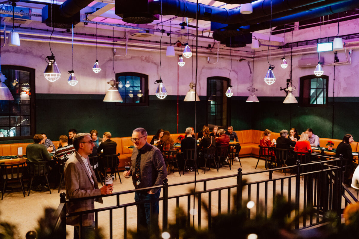 wigilia firmowa spoldzielnia koneser fotograf na impreze 018 Ludzie siedzą przy stolikach i rozmawiają w nowoczesnej restauracji lub kawiarni w stylu industrialnym z ciepłym oświetleniem. Dwóch mężczyzn stoi rozmawiając na pierwszym planie, podczas gdy inni cieszą się jedzeniem i napojami w grupach w tle.