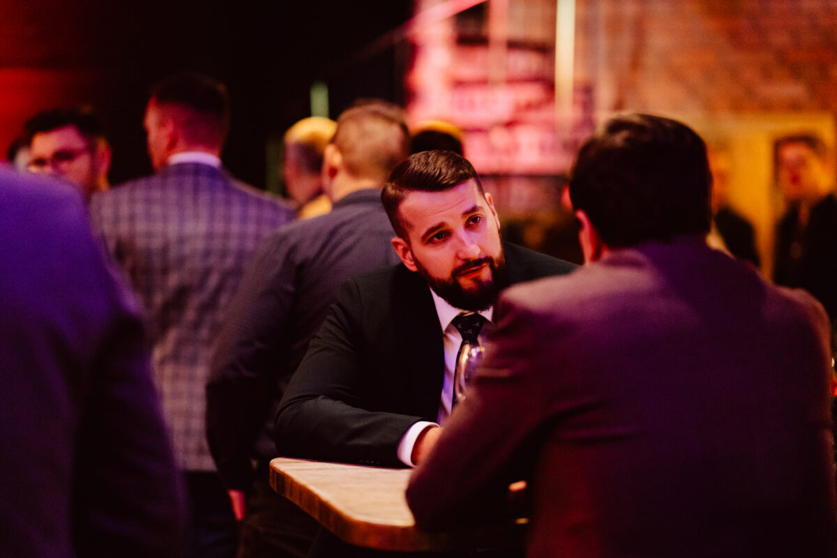 wigilia firmowa spoldzielnia koneser fotograf na impreze 019 Mężczyzna z brodą w garniturze siedzi przy stole, słuchając innej osoby na zatłoczonym, słabo oświetlonym spotkaniu towarzyskim z ludźmi w tle.