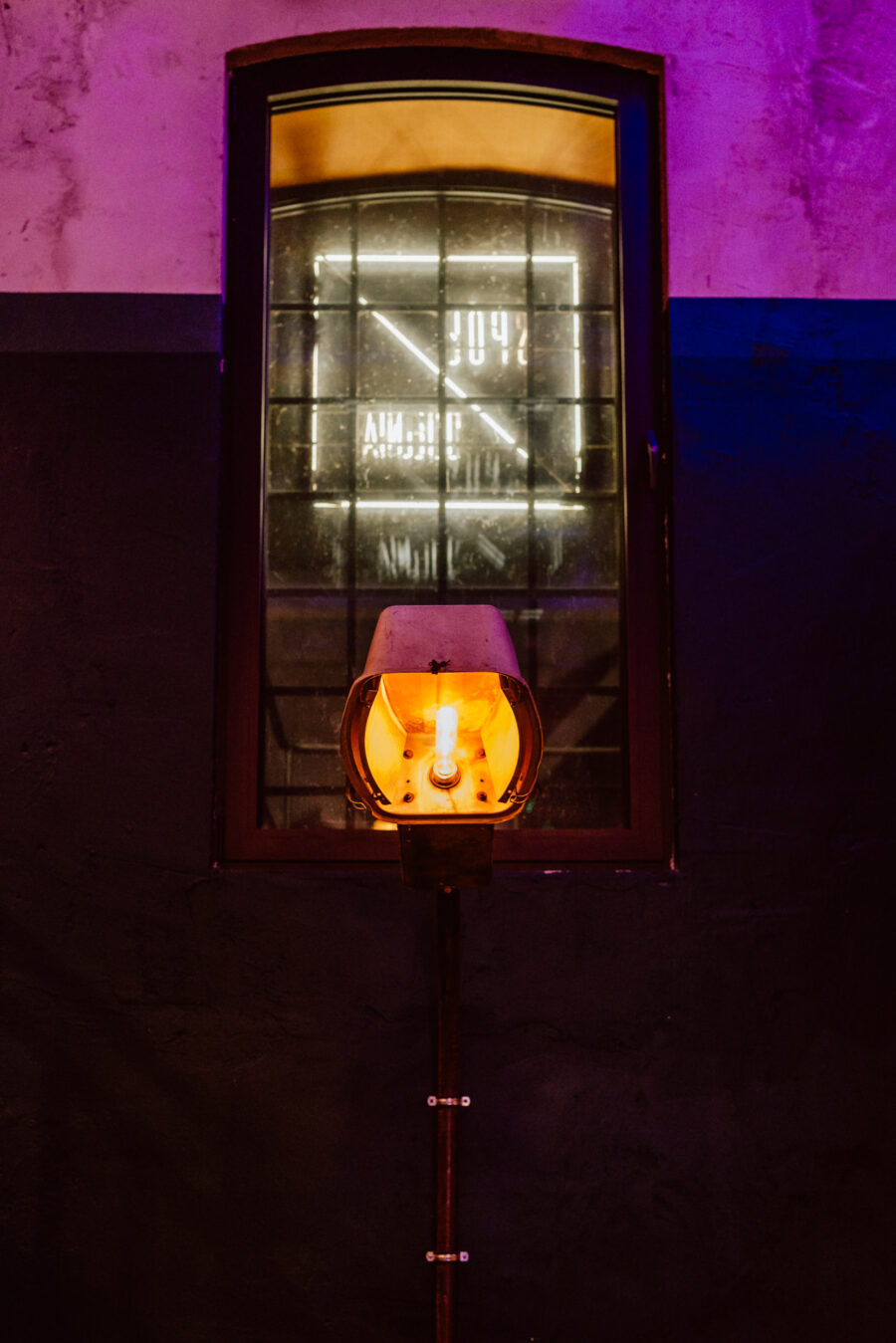 wigilia firmowa spoldzielnia koneser fotograf na impreze 020 Lampa w stylu vintage z odsłoniętą żarówką świeci ciepło przed oknem. Okno odbija neony i geometryczne kształty z zewnątrz, dodając kolorowe światło do przyciemnionego pokoju.