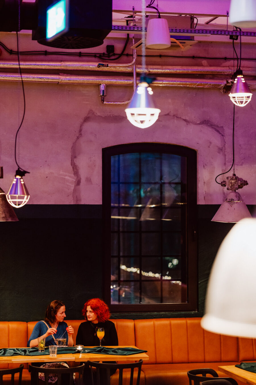 wigilia firmowa spoldzielnia koneser fotograf na impreze 021 Dwie kobiety siedzą na opalonej kanapie w nowoczesnym, słabo oświetlonym barze, rozmawiając przy drinku. Odsłonięte rury, wiszące światła i duże okno z nocnymi światłami miasta dodają przytulnej atmosfery.
