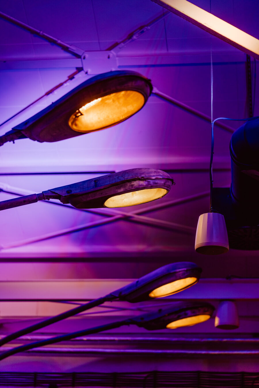 wigilia firmowa spoldzielnia koneser fotograf na impreze 022 Cztery latarnie uliczne są zamontowane wewnątrz budynku z fioletową i pomarańczową poświatą, tworząc futurystyczną i artystyczną atmosferę. Wśród świateł wisi biała cylindryczna oprawa. Sufit ma geometryczne wzory.