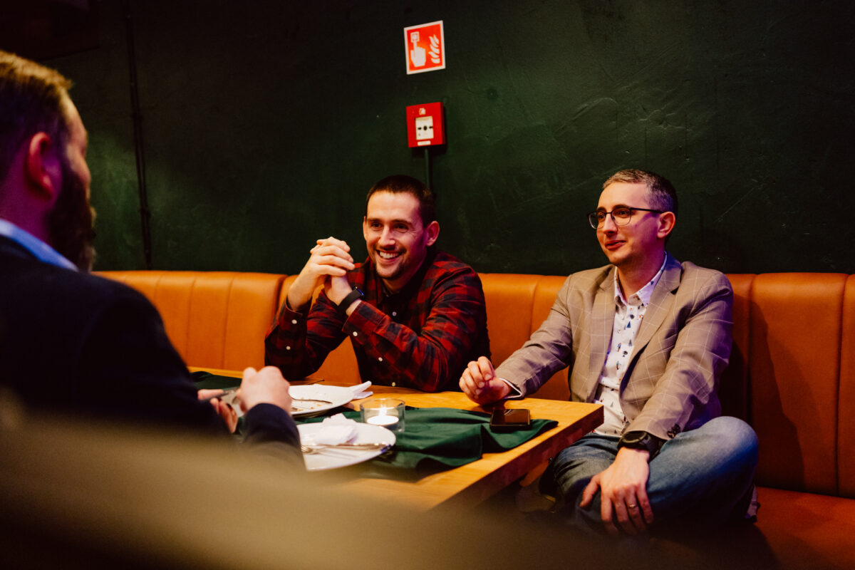 wigilia firmowa spoldzielnia koneser fotograf na impreze 024 Trzech mężczyzn siedzi przy stoliku w restauracji i prowadzi rozmowę. Dwóch mężczyzn stoi twarzą do kamery, jeden uśmiecha się i ściska dłonie, drugi ma na sobie okulary i marynarkę. Trzeci mężczyzna stoi tyłem do kamery.