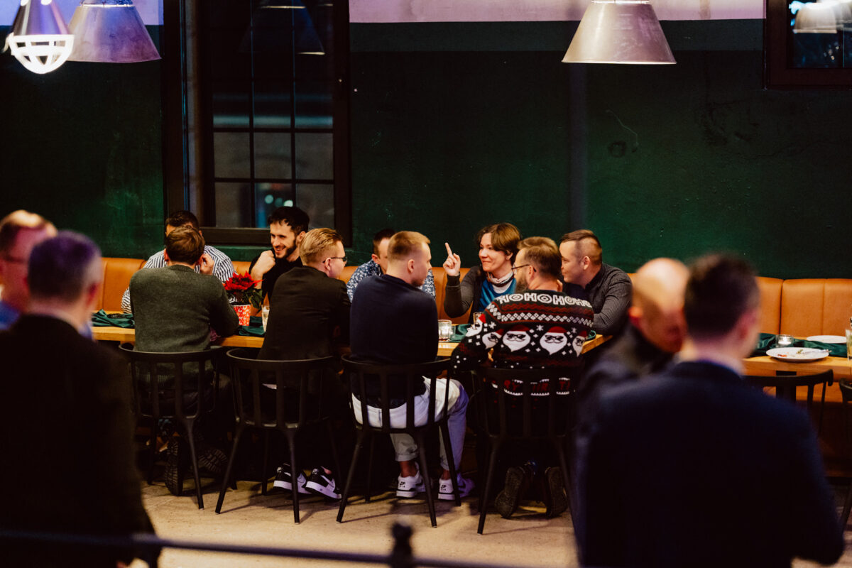 wigilia firmowa spoldzielnia koneser fotograf na impreze 025 Grupa ludzi siedzi wokół długiego stołu w restauracji, zaangażowana w rozmowę i uśmiechnięta. Otoczenie jest ciepło oświetlone, z ciemnozielonymi ścianami i nowoczesnymi wiszącymi lampami nad nimi.