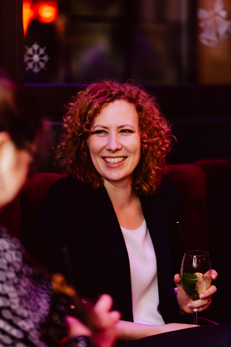 wigilia firmowa spoldzielnia koneser fotograf na impreze 026 Kobieta z kręconymi rudymi włosami uśmiecha się, siedząc w pomieszczeniu, trzymając w dłoni drinka z miętą i lodem. Tło jest delikatnie oświetlone ciepłymi kolorami, a na oknie widoczna jest dekoracja z płatków śniegu.