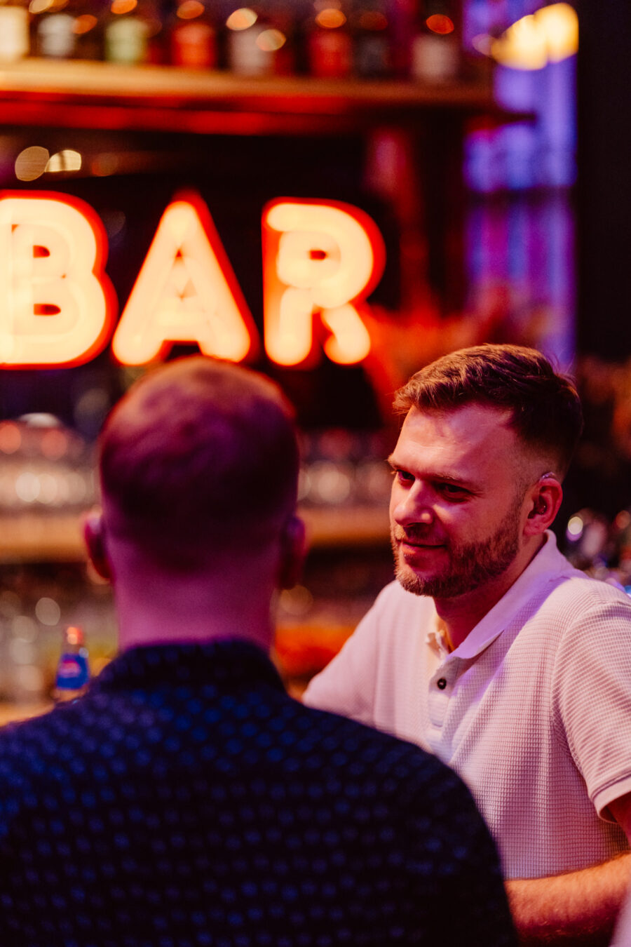 wigilia firmowa spoldzielnia koneser fotograf na impreze 027 Dwóch mężczyzn siedzi przy barze i rozmawia. Jeden z nich, zwrócony w stronę kamery, ma na sobie białą koszulę i uśmiecha się, podczas gdy drugi jest odwrócony plecami do kamery. W tle świeci neonowy napis "BAR".