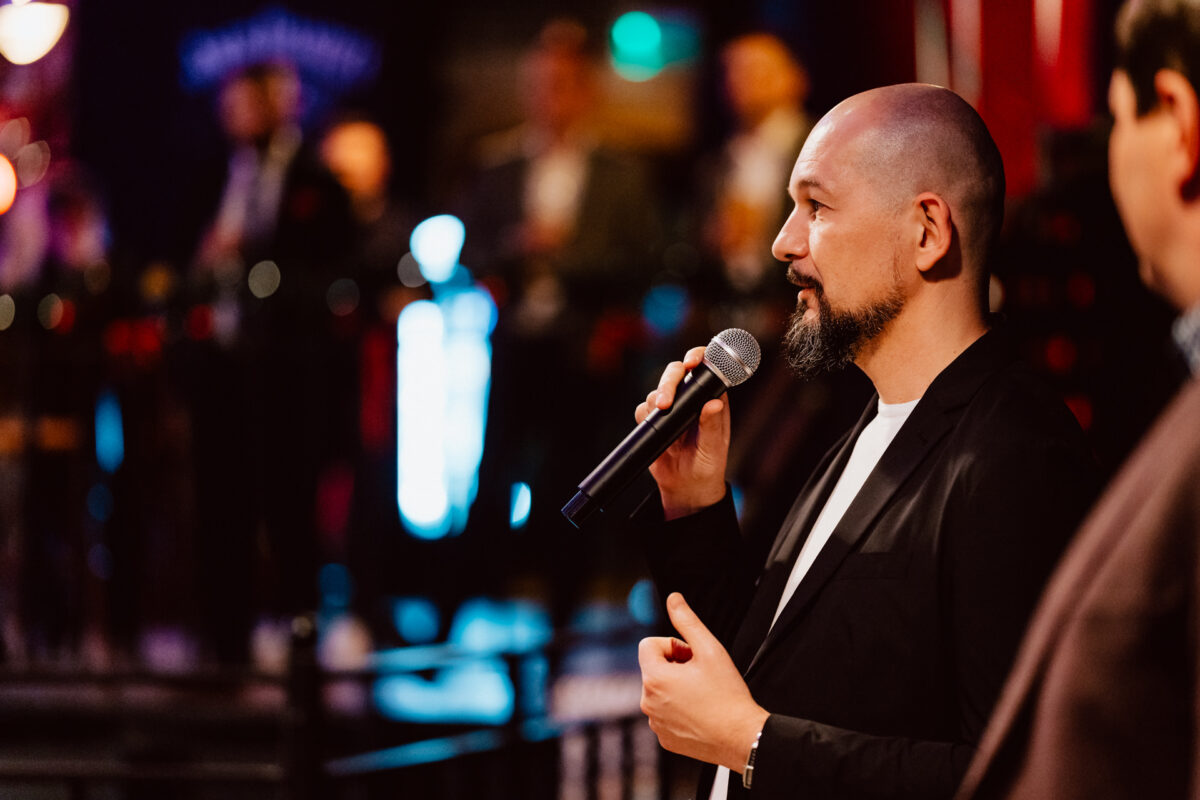 wigilia firmowa spoldzielnia koneser fotograf na impreze 028 Mężczyzna z ogoloną głową i brodą, ubrany w czarną kurtkę, mówi do mikrofonu podczas imprezy w pomieszczeniu. Tło jest rozmyte kolorowymi światłami i stojącymi ludźmi.