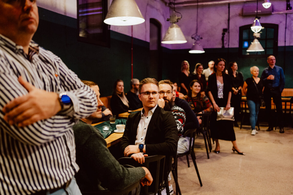 wigilia firmowa spoldzielnia koneser fotograf na impreze 029 Grupa ludzi siedzi i stoi w ciepło oświetlonym pomieszczeniu, uważnie obserwując coś poza kamerą. Niektórzy są ubrani swobodnie, podczas gdy inni noszą stroje biznesowe. Atmosfera jest swobodna i towarzyska.