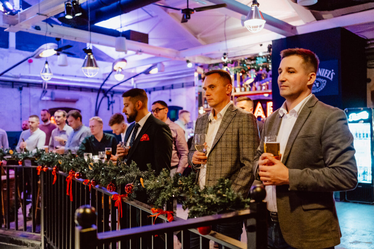 wigilia firmowa spoldzielnia koneser fotograf na impreze 030 Grupa dobrze ubranych ludzi stoi w barze udekorowanym świątecznymi girlandami i światełkami, trzymając drinki i uważnie obserwując coś poza kamerą w świątecznej, ciepło oświetlonej atmosferze.