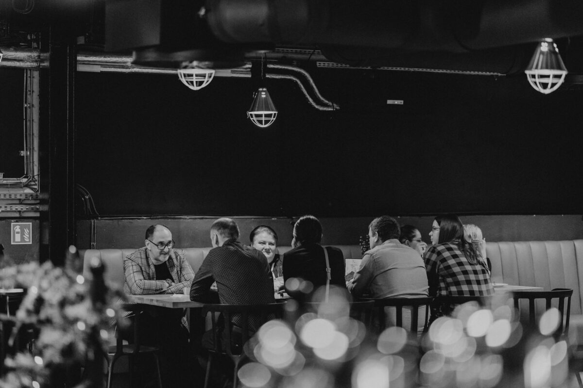 wigilia firmowa spoldzielnia koneser fotograf na impreze 031 Grupa ludzi siedzi przy stole w słabo oświetlonej restauracji lub barze i prowadzi rozmowę. Obraz jest czarno-biały, z rozmytymi światłami na pierwszym planie i oświetleniem przemysłowym nad głową.