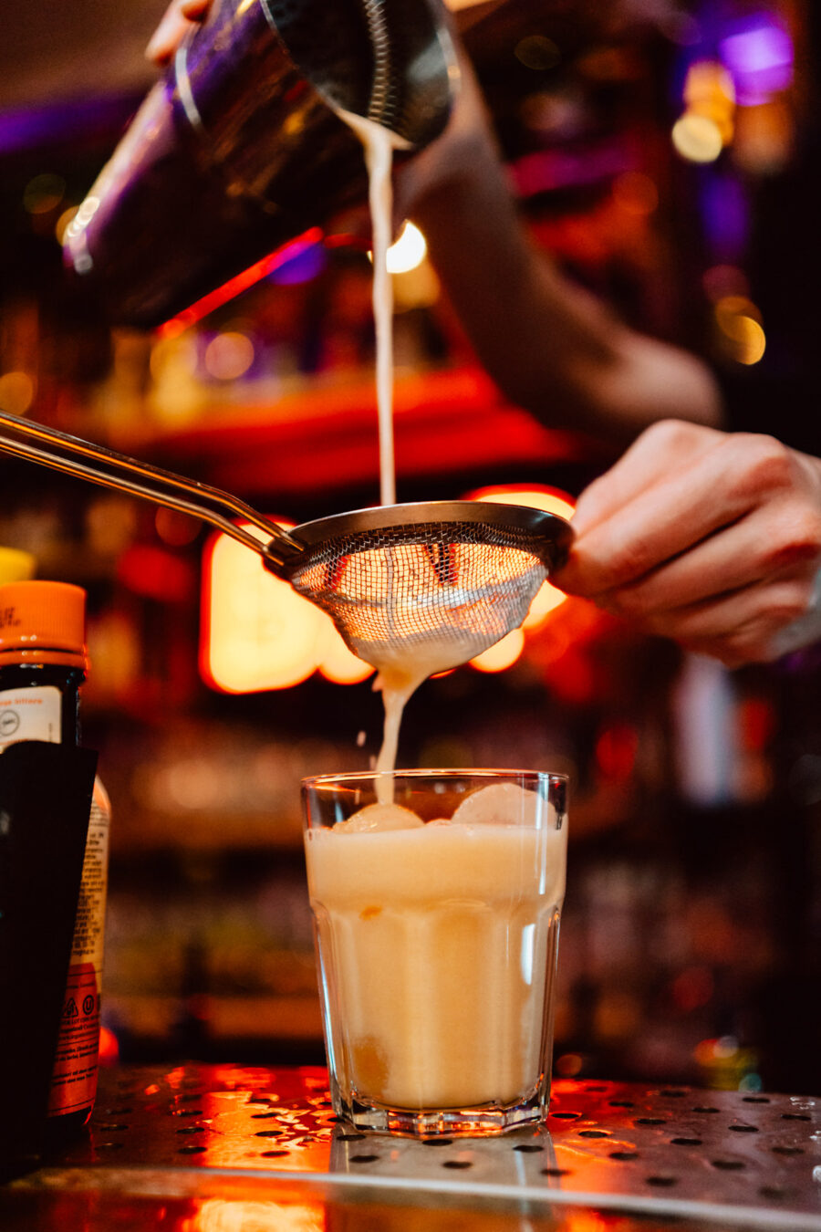 wigilia firmowa spoldzielnia koneser fotograf na impreze 032 Barman nalewa kremowy koktajl przez sitko do szklanki wypełnionej kostkami lodu w słabo oświetlonym barze, z kolorowymi światłami rozmytymi w tle.