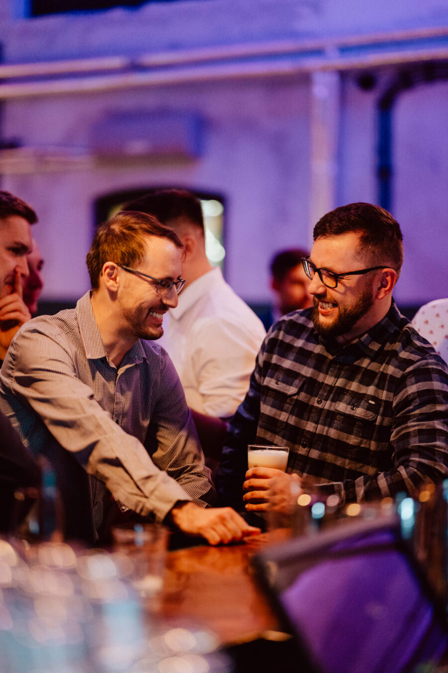 wigilia firmowa spoldzielnia koneser fotograf na impreze 033 Dwóch mężczyzn stoi przy barze, uśmiechając się i rozmawiając. Jeden z nich trzyma szklankę piwa, a obaj mają na sobie okulary i swobodne koszule. Tło jest delikatnie oświetlone fioletowym odcieniem, a w pobliżu widoczne są inne osoby.