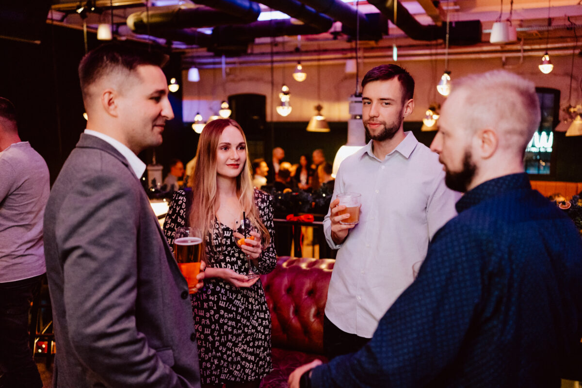 wigilia firmowa spoldzielnia koneser fotograf na impreze 034 Cztery osoby stoją razem na imprezie towarzyskiej, trzymając drinki i rozmawiając. Trzech mężczyzn i jedna kobieta są zaangażowani w rozmowę, z żywym, ciepło oświetlonym tłem i gośćmi widocznymi za nimi.