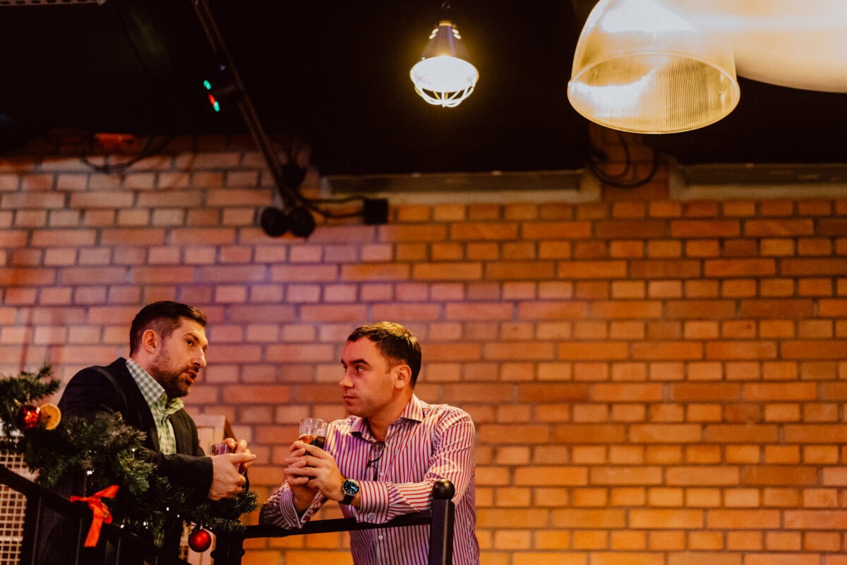 wigilia firmowa spoldzielnia koneser fotograf na impreze 035 Dwóch mężczyzn stoi przy ceglanej ścianie i rozmawia, trzymając drinki. Na balustradzie przed nimi widoczne są świąteczne dekoracje z ornamentami i girlandami. Ciepłe oświetlenie tworzy przytulną atmosferę.