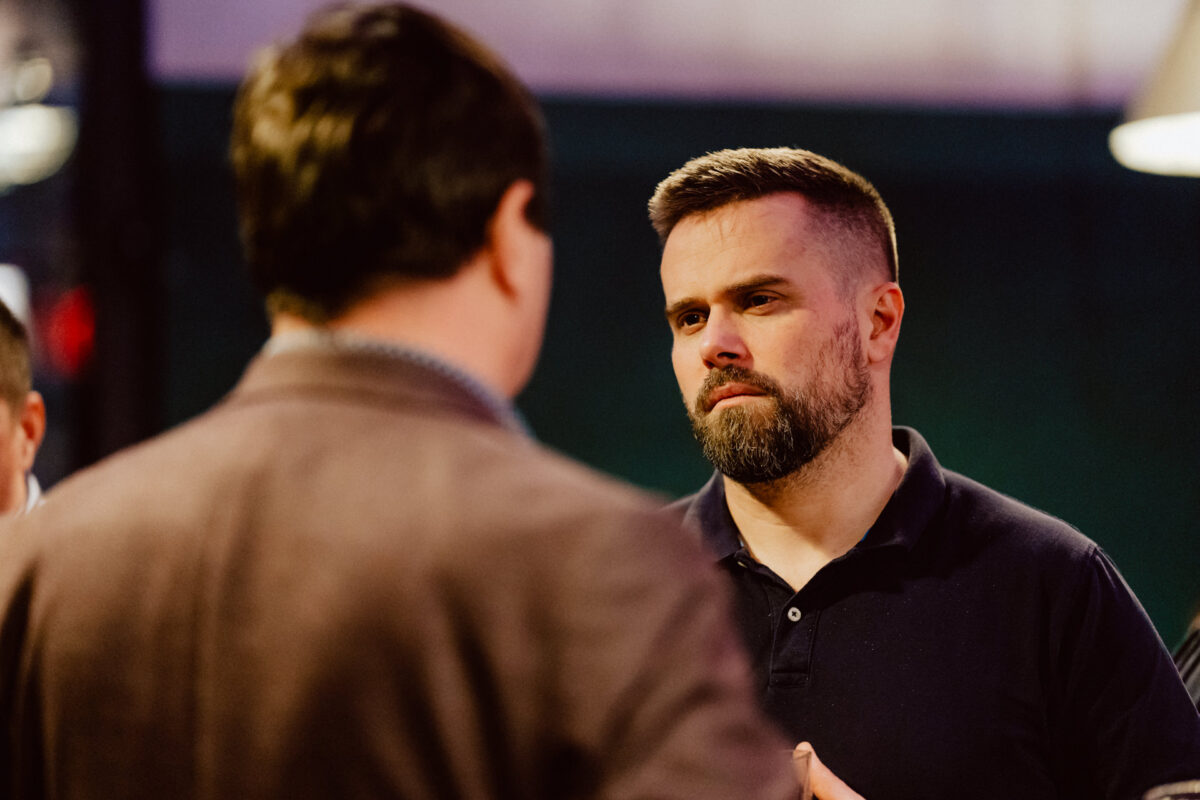 wigilia firmowa spoldzielnia koneser fotograf na impreze 037 Brodaty mężczyzna w czarnej koszulce polo słucha uważnie innej osoby odwróconej plecami do kamery podczas rozmowy w pomieszczeniu. Tło jest delikatnie rozmyte.